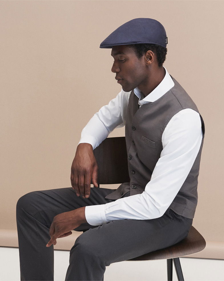 Mann sitzt seitlich auf Stuhl, trägt weiße Bluse, graue Weste, dunkle Hose und navyblaue Mütze, schaut nach unten.