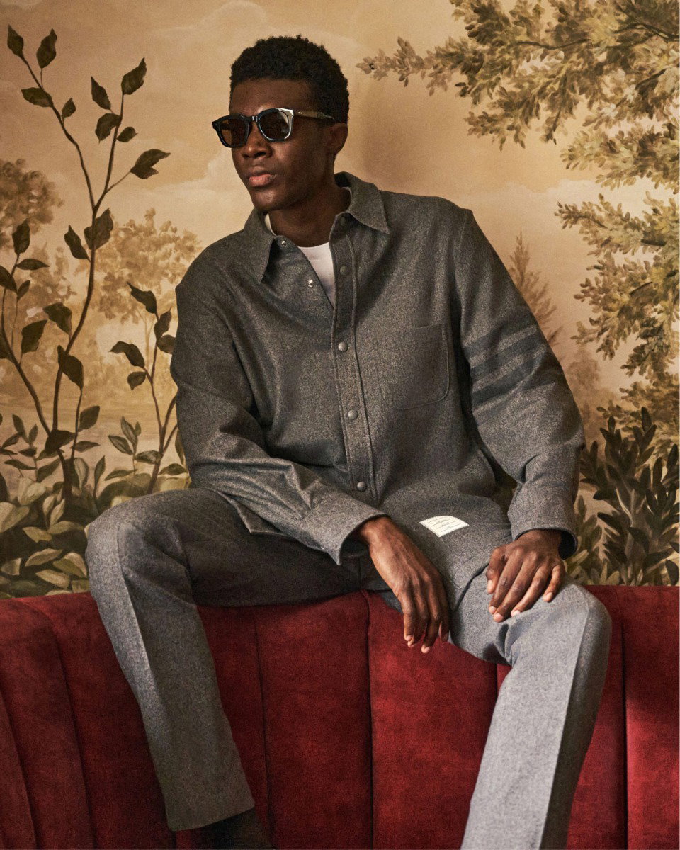 Mann mit Sonnenbrille in grauem Overshirt und Hose sitzt lässig auf rotem Sofa vor gemusterter Tapete mit Pflanzenmotiven.