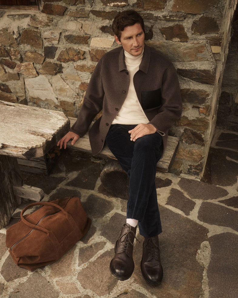 Mann sitzt auf Holzbänkchen vor Steinmauer, trägt braune Jacke, cremefarbenen Pullover, dunkelblaue Cordhose, braune Schnürboots.