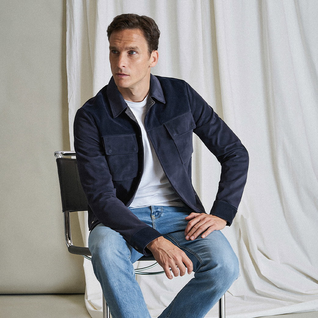 Mann sitzt auf Stuhl, trägt weiße Shirt, dunkelblaue Jacke und helle Jeans, schaut nach links, vor weißem Vorhang.