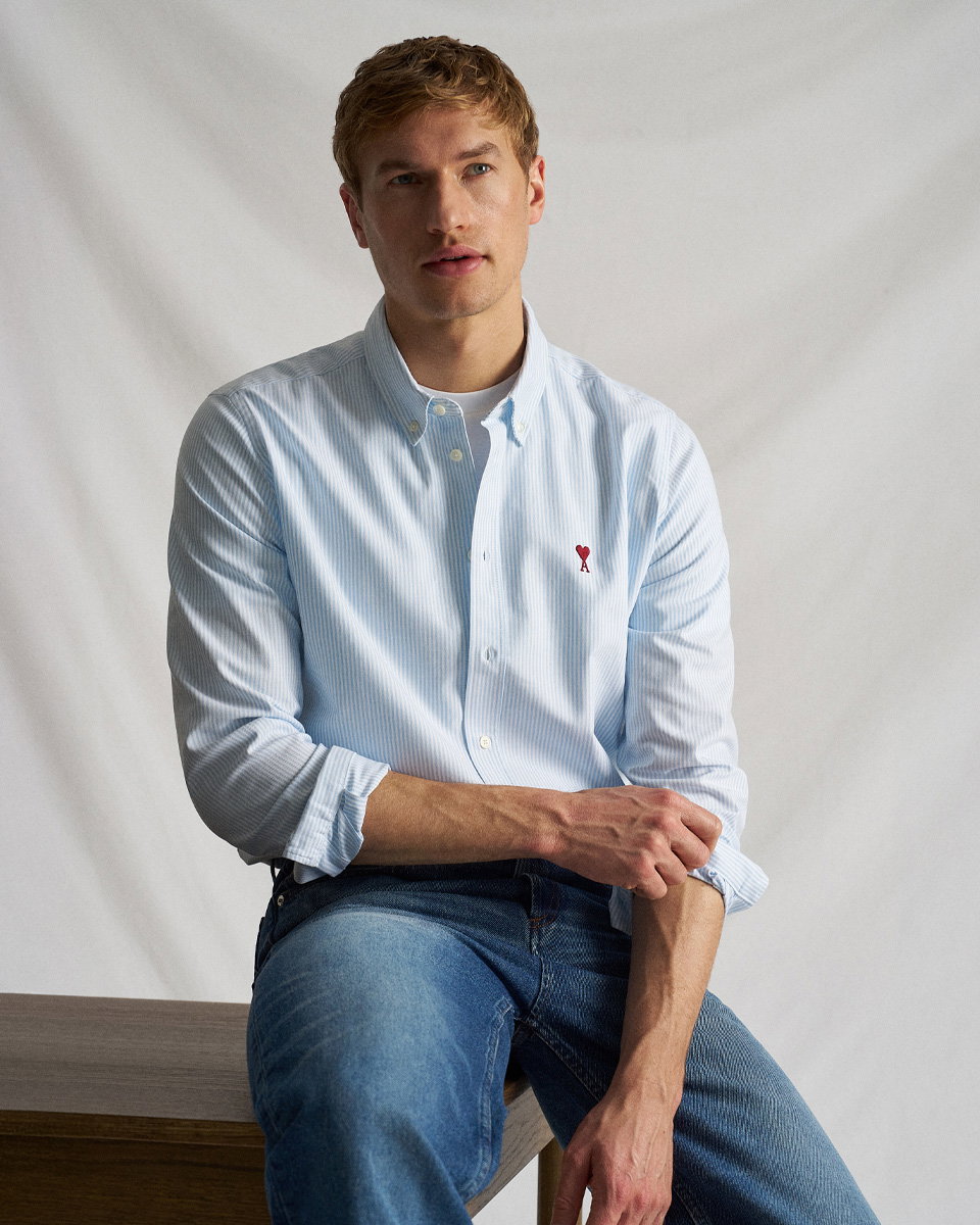 Mann sitzt auf Tisch, trägt hellblau-weiß gestreiftes Hemd mit rotem Herz-Logo und blaue Jeans, schaut nach rechts.