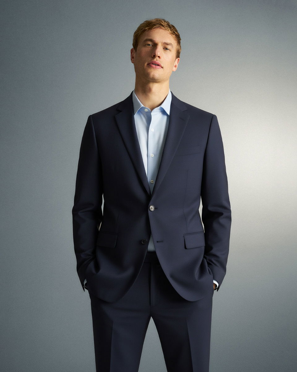 Mann steht mit Händen in Hosentaschen, trägt navyblauen Anzug und hellblaues Hemd, neutraler grauer Hintergrund.