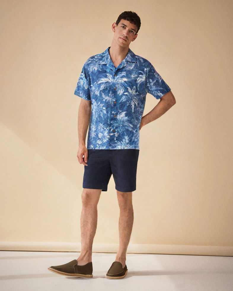 Junger Mann steht vor beigem Hintergrund, trägt blau-weiß gemustertes Kurzarmhemd, navy Shorts und olivgrüne Espadrilles.