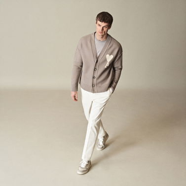 Herrenportrait: Mann trägt cremefarbene Hose, hellgraues Shirt, beigen Cardigan mit Herzmotiv und weiße Sneakers, vor neutralem Hintergrund.