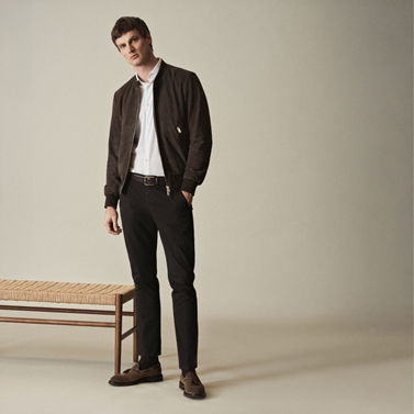 Mann trägt schwarze Hose, weißes Hemd, dunkelbraunen Veloursleder-Blouson und braune Loafer, steht neben Holzbank.