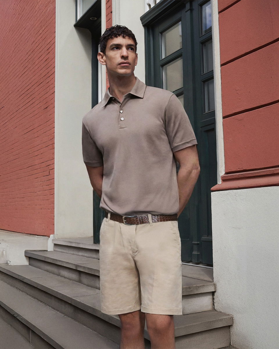 Mann steht vor Haustür, trägt hellbraunes Poloshirt, beige Chinoshorts und braunen geflochtenen Gürtel.