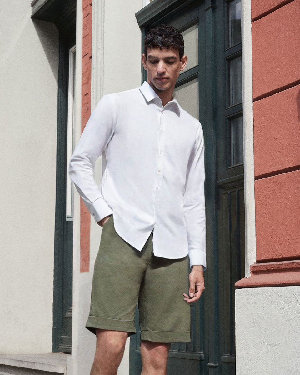 Mann steht vor Hauswand mit roten und weißen Fassadenteilen, trägt weißes Hemd und olivgrüne Shorts, Blick nach unten.