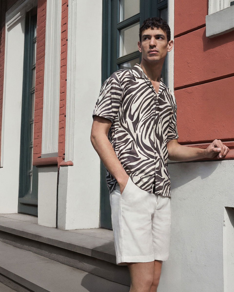 Mann lehnt an roter Hauswand, trägt weiß-braunes Zebra-Muster Kurzarmhemd und weiße Shorts, schaut nach rechts.