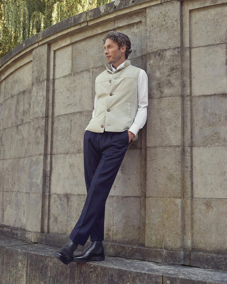 Mann lehnt an grauer Steinmauer, trägt weiße Weste, weißes Hemd, blaue Hose und schwarze Boots, Blick nach rechts.