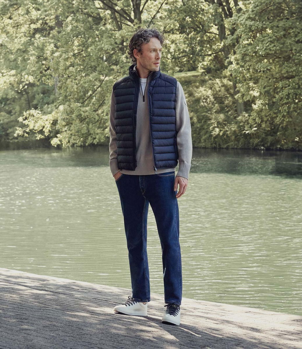 Mann mit blauer Weste, beigem Pullover, blauer Jeans und weißen Sneakern steht am Seeufer vor grünem Wald im Sonnenlicht.