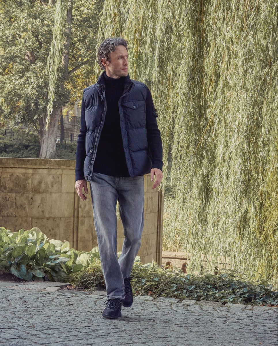 Mann mit blauem Rollkragenpullover, blauer Weste und Jeans läuft auf gepflastertem Weg vor See und Weidenbaum.