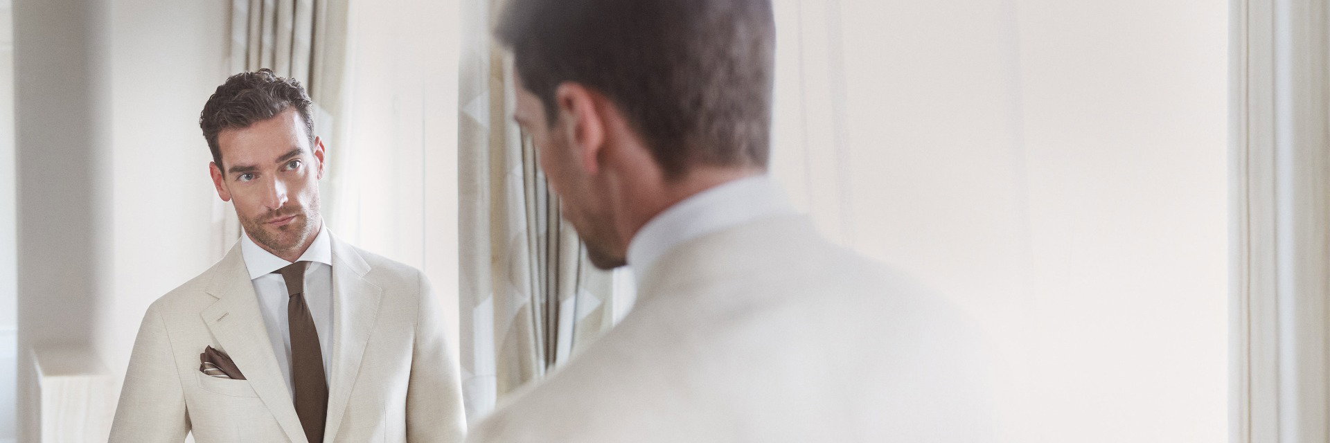 Mann im cremefarbenen Sakko, weißem Hemd, brauner Krawatte und Einstecktuch blickt ernst in einen Spiegel. Mann im cremefarbenen Sakko, weißem Hemd, brauner Krawatte und Einstecktuch blickt ernst in einen Spiegel.