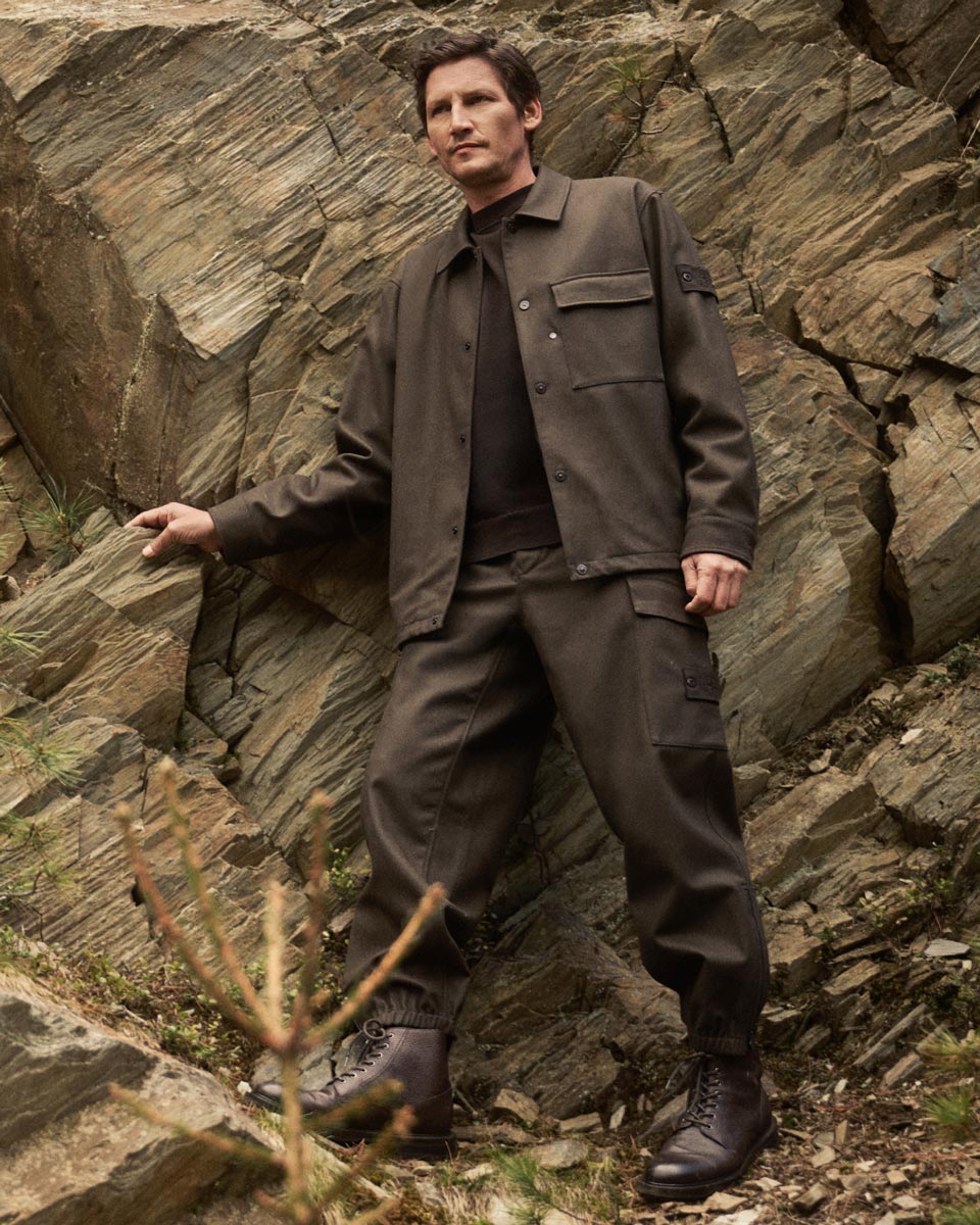 Mann steht vor Felswand, trägt braunes Overshirt, Cargo-Hose, braune Schnürstiefel, Naturkulisse mit Felsen und Pflanzen. Mann steht vor Felswand, trägt braunes Overshirt, Cargo-Hose, braune Schnürstiefel, Naturkulisse mit Felsen und Pflanzen.