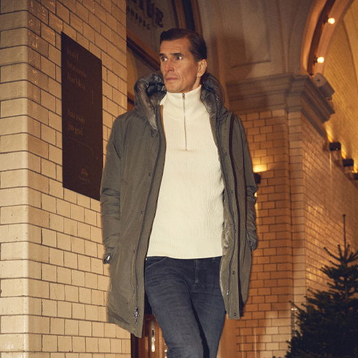 Mann in weißem Troyer, grüner Parka und dunkler Jeans lehnt an beleuchteter Steinwand in urbanem Umfeld bei Abendlicht. Mann in weißem Troyer, grüner Parka und dunkler Jeans lehnt an beleuchteter Steinwand in urbanem Umfeld bei Abendlicht.