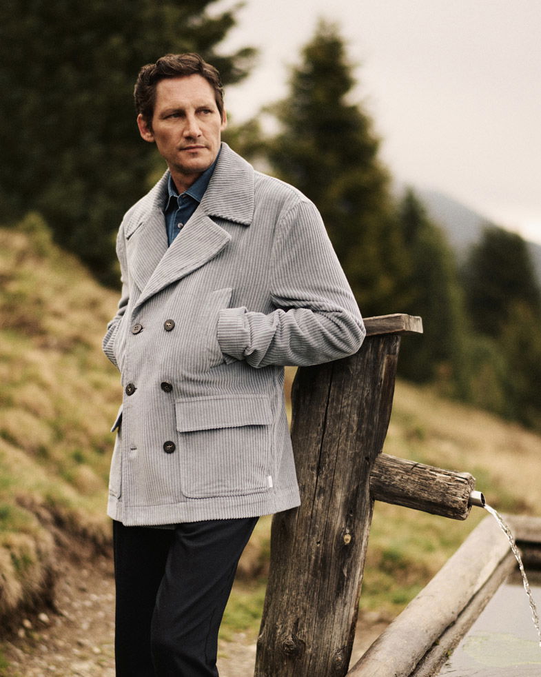 Mann in rauchblauer Cordjacke und blauem Jeanshemd lehnt an Holzbrunnen vor bewaldeter Berglandschaft.