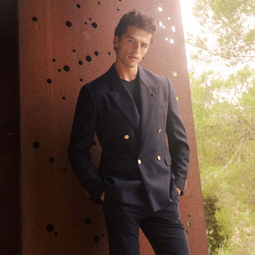 Mann lehnt an rostiger Wand, trägt navy zweireihiges Sakko mit goldenen Knöpfen, passendes Shirt und Hose, Outdoor-Szenerie.