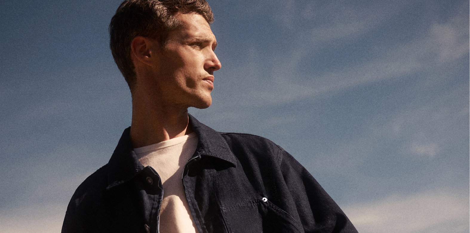 Mann mit kurzem braunem Haar trägt weißes T-Shirt und navyblaues Overshirt, blickt rechts, blauer Himmel mit Wolken im Hintergrund.