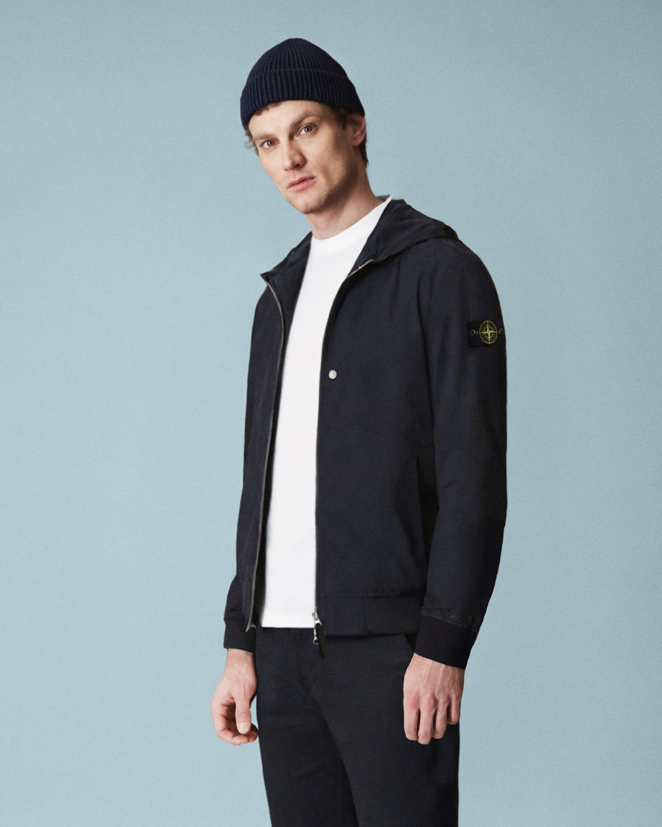 Mann vor blauem Hintergrund trägt schwarze Strickmütze, schwarze Jacke mit Logo am Arm, weißes Shirt und schwarze Hose. Mann vor blauem Hintergrund trägt schwarze Strickmütze, schwarze Jacke mit Logo am Arm, weißes Shirt und schwarze Hose.