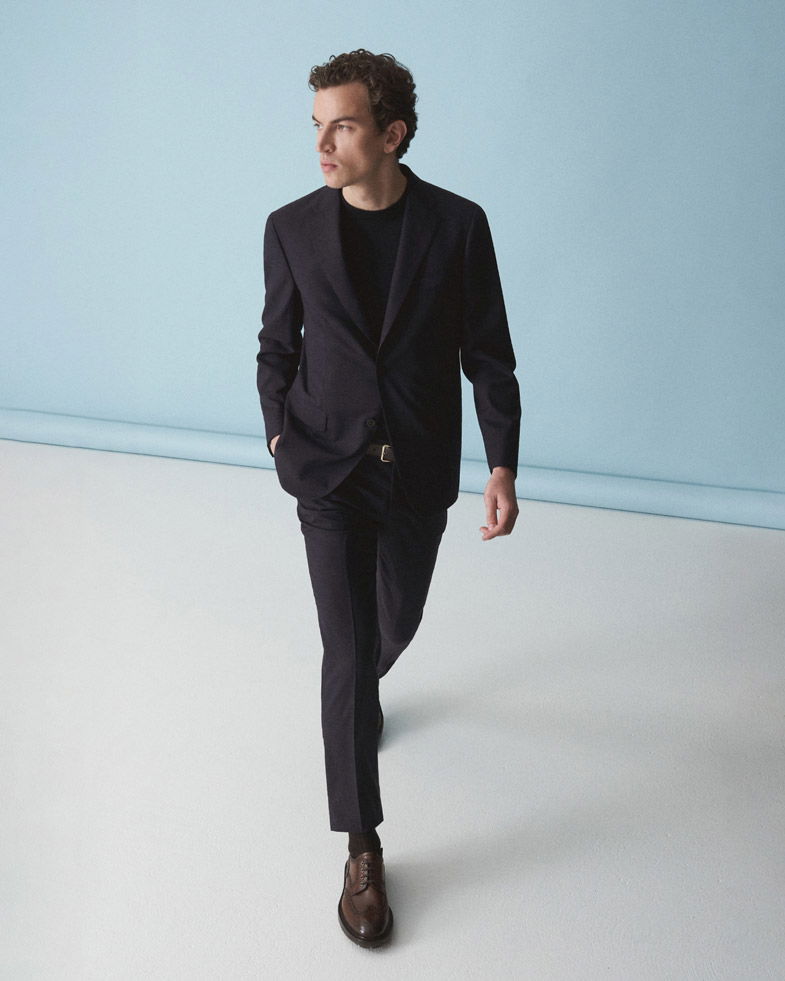 Junger Mann in dunkelblauem Anzug und schwarzem Shirt geht vor hellblauer Wand, trägt braune Lederschnürschuhe.