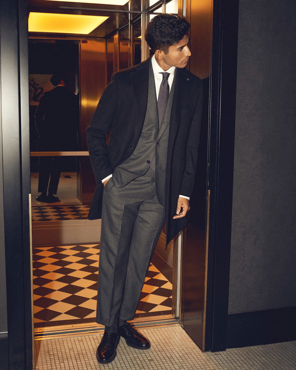 Mann in grauem Anzug, weißem Hemd, schwarzer Krawatte und schwarzem Mantel lehnt im geöffneten Fahrstuhl an der Tür. Mann in grauem Anzug, weißem Hemd, schwarzer Krawatte und schwarzem Mantel lehnt im geöffneten Fahrstuhl an der Tür.