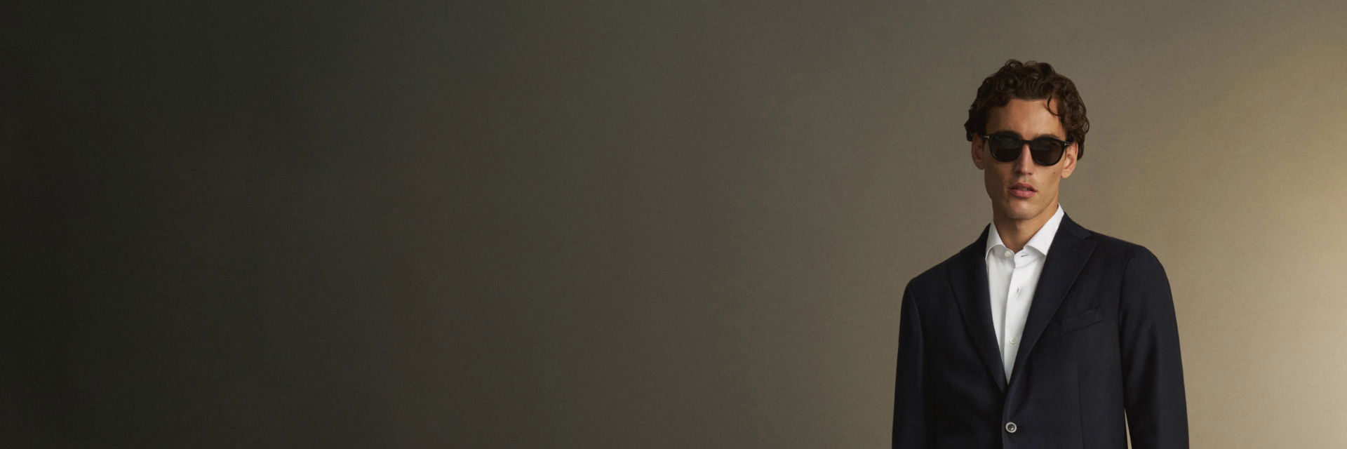 Mann mit kurzen lockigen Haaren trägt navy Sakko, weißes Hemd und schwarze Sonnenbrille vor dunklem neutralem Hintergrund. Mann mit kurzen lockigen Haaren trägt navy Sakko, weißes Hemd und schwarze Sonnenbrille vor dunklem neutralem Hintergrund.