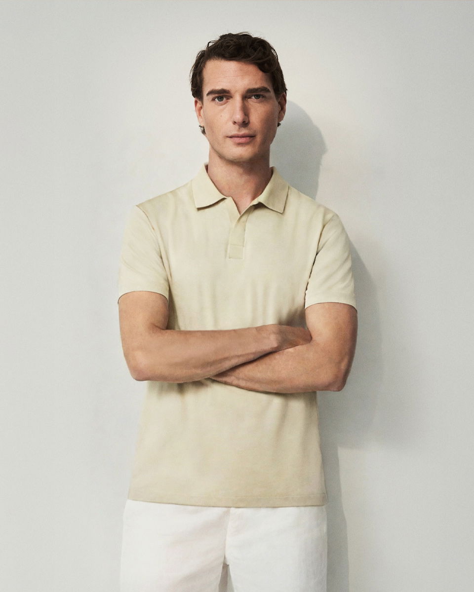 Mann steht vor weißer Wand, trägt beige Poloshirt und weiße Hose, Arme verschränkt, schaut gerade in die Kamera.