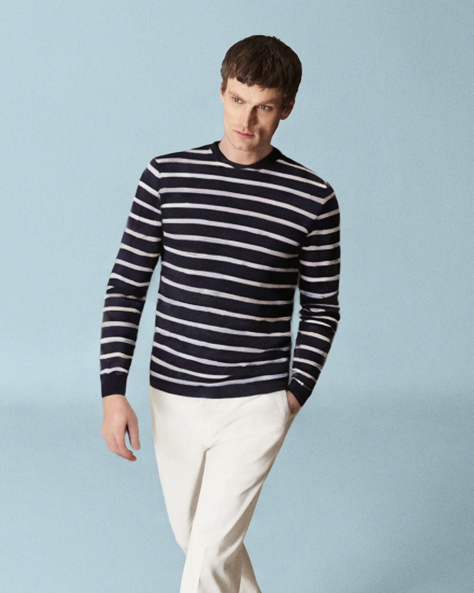 Mann vor hellblauem Hintergrund trägt weißen gestreiften Pullover und weiße Hose, eine Hand in der Tasche, Blick nach rechts. Mann vor hellblauem Hintergrund trägt weißen gestreiften Pullover und weiße Hose, eine Hand in der Tasche, Blick nach rechts.