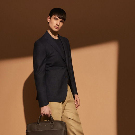 Junger Mann vor hellbrauner Wand trägt dunkelblaues Sakko, schwarzes Shirt, beige Hose und hält braune Aktentasche in der Hand.