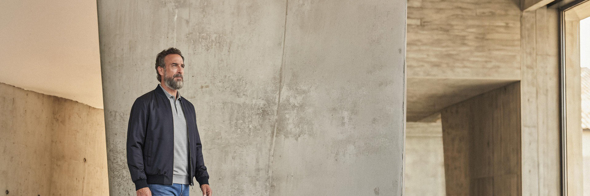 Mann mit grauem Poloshirt, dunkelblauer Jacke und blauer Jeans steht vor heller roher Betonwand in modernem Raum. Mann mit grauem Poloshirt, dunkelblauer Jacke und blauer Jeans steht vor heller roher Betonwand in modernem Raum.