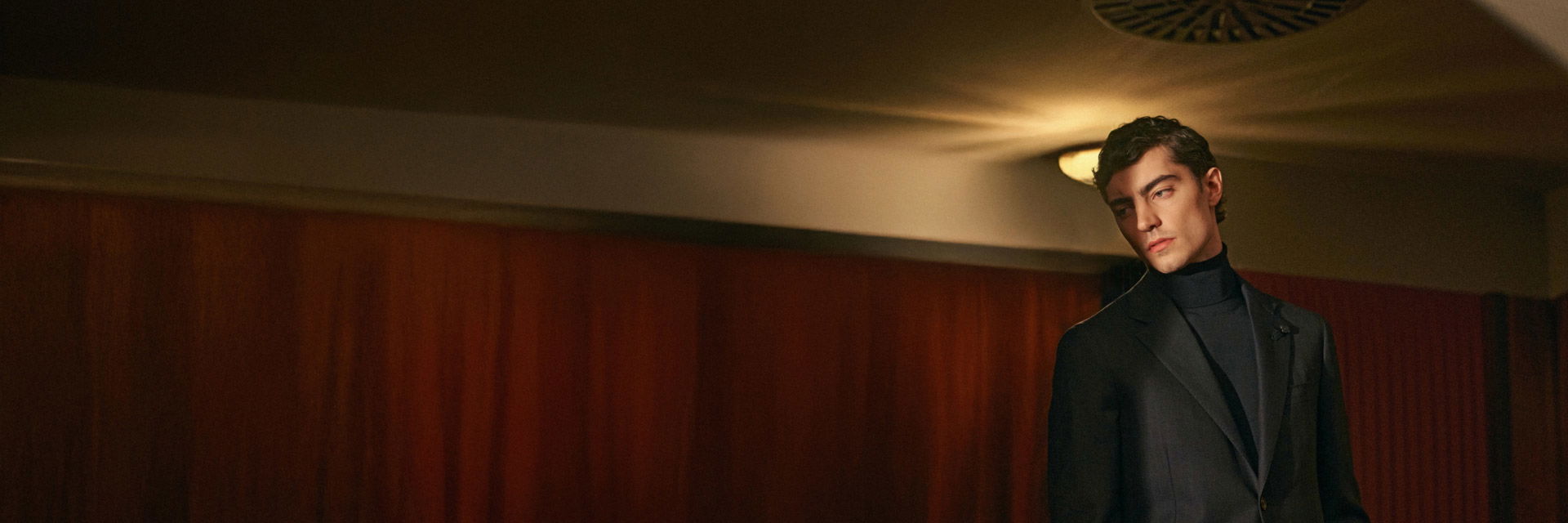 Mann steht vor rotem Vorhang, trägt dunkles Sakko über dunkelgrauem Rollkragenpullover, Blick leicht seitlich, gedämpftes Licht. Mann steht vor rotem Vorhang, trägt dunkles Sakko über dunkelgrauem Rollkragenpullover, Blick leicht seitlich, gedämpftes Licht.