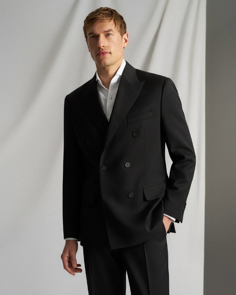 Mann mit blondem Haar trägt schwarzen, doppelreihigen Anzug und weißes Hemd, steht vor weißem Hintergrund.