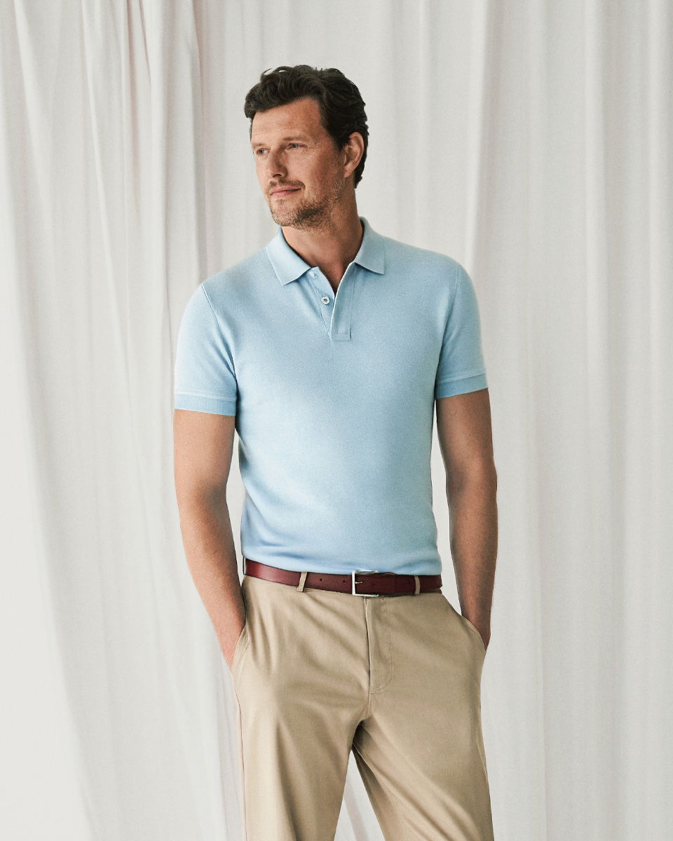 Mann steht vor weißem Vorhang, trägt hellblaues Poloshirt, beige Hose und braunen Gürtel, Hände in Taschen.