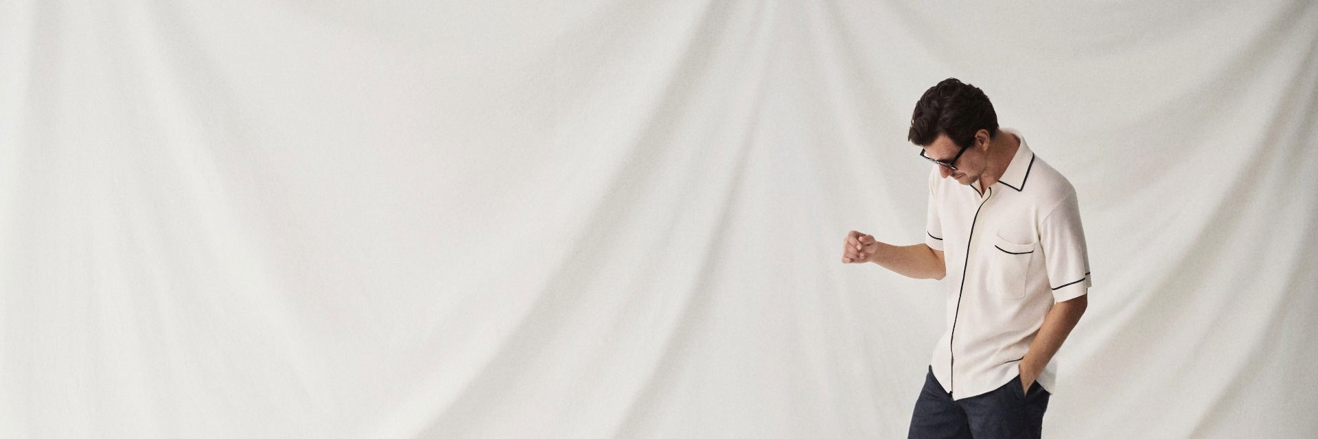 Mann mit braunen Haaren trägt weißes Kurzarmhemd mit schwarzen Details und dunkle Hose vor weißem Stoffhintergrund. Mann mit braunen Haaren trägt weißes Kurzarmhemd mit schwarzen Details und dunkle Hose vor weißem Stoffhintergrund.