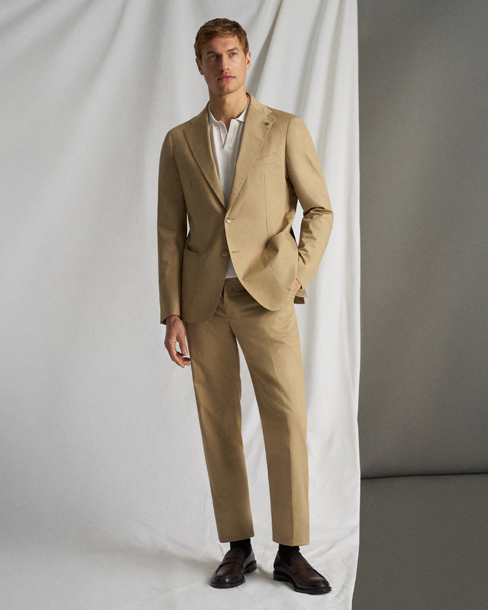 Beiger Anzug mit weißem Poloshirt, dunkelbraunen Loafern, stehend vor weißem Vorhang, linker Hand in Hosentasche.