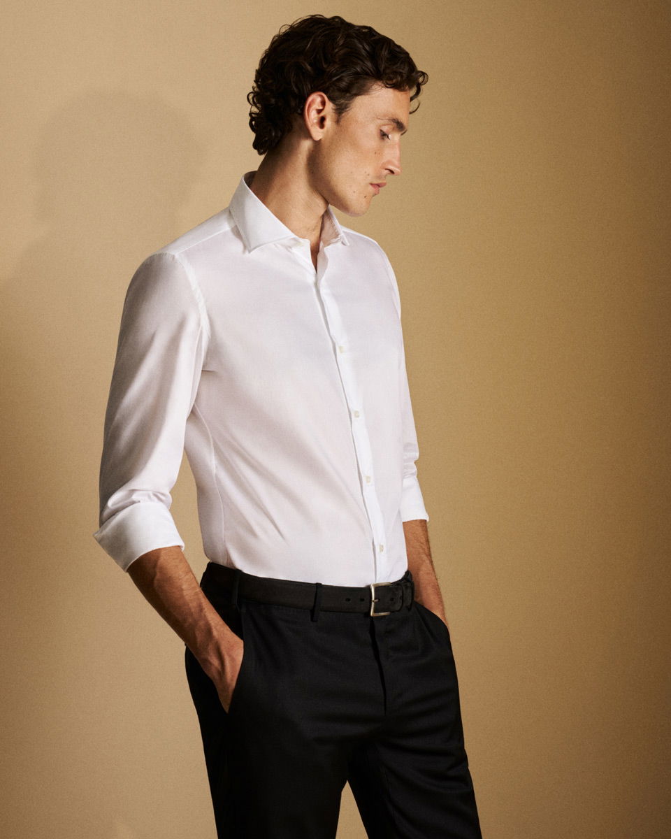 Mann im weißen Hemd mit gekrempelten Ärmeln und schwarzer Hose, der seitlich mit Händen in den Taschen steht.