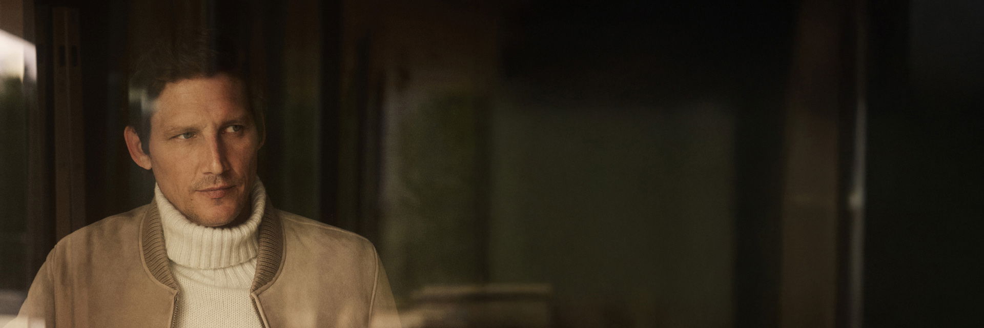 Mann mit beiger Lederjacke und weißem Rollkragenpullover schaut nachdenklich zur Seite, dunkler Hintergrund. Mann mit beiger Lederjacke und weißem Rollkragenpullover schaut nachdenklich zur Seite, dunkler Hintergrund.