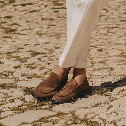 Braune Herren-Loafer aus Wildleder getragen mit cremefarbener, umgekrempelter Hose auf Kopfsteinpflaster. Braune Herren-Loafer aus Wildleder getragen mit cremefarbener, umgekrempelter Hose auf Kopfsteinpflaster.