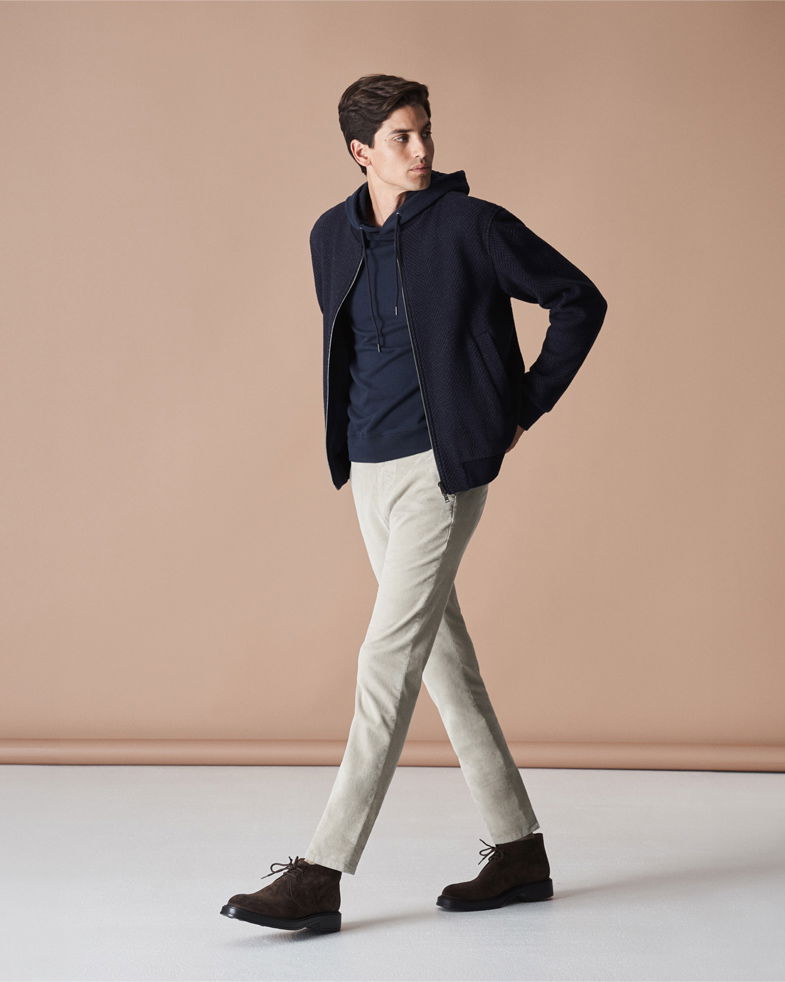 Mann trägt navy Bomberjacke, dunkelblauen Hoodie, beige Hose und braune Schnürboots vor beige Hintergrund. Mann trägt navy Bomberjacke, dunkelblauen Hoodie, beige Hose und braune Schnürboots vor beige Hintergrund.