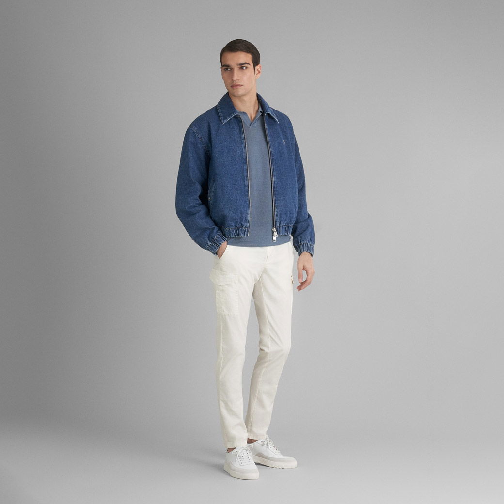 Mann trägt blaue Jeans-Blousonjacke, graublaues Strickpolo, offwhite Cargohose und weiße Sneaker vor grauem Hintergrund.