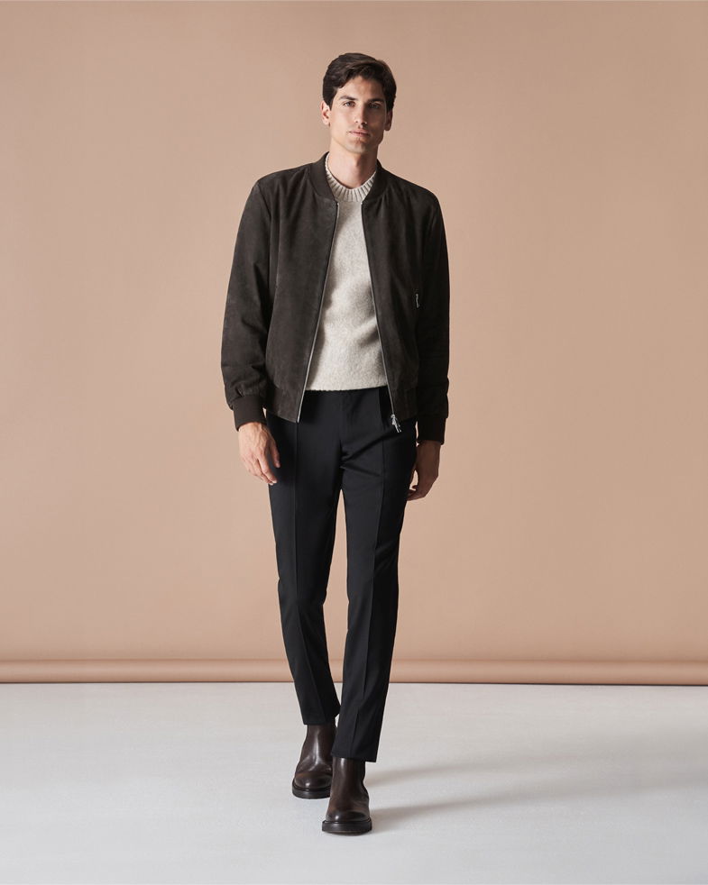 Mann trägt braune Veloursleder-Bomberjacke, beigen Pullover, schwarze Hose und braune Stiefel, vor beigem Hintergrund. Mann trägt braune Veloursleder-Bomberjacke, beigen Pullover, schwarze Hose und braune Stiefel, vor beigem Hintergrund.