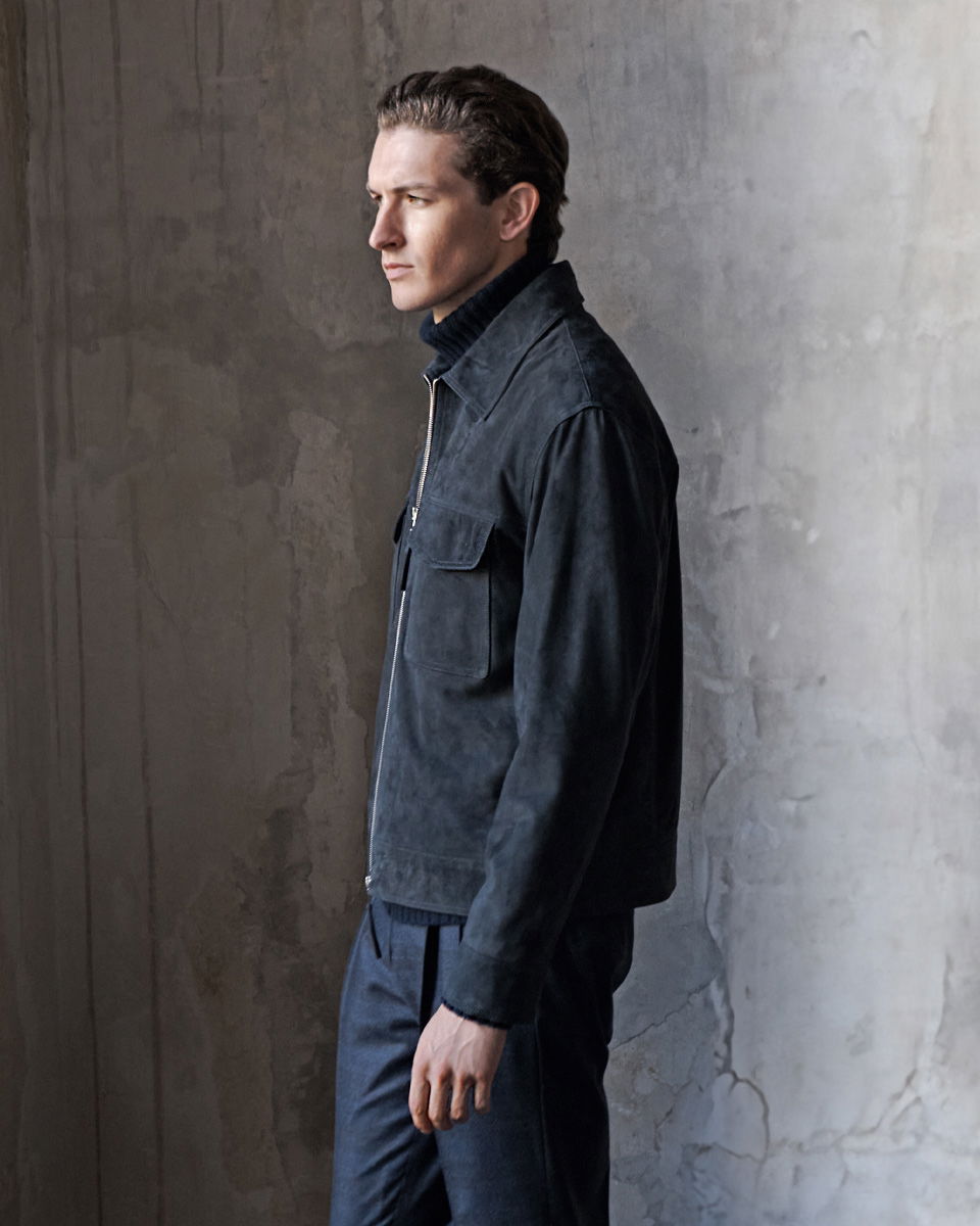 Mann im Profil trägt navy Velourslederjacke, dunkle Hose und schwarzen Rollkragenpullover vor grauer Wand.
