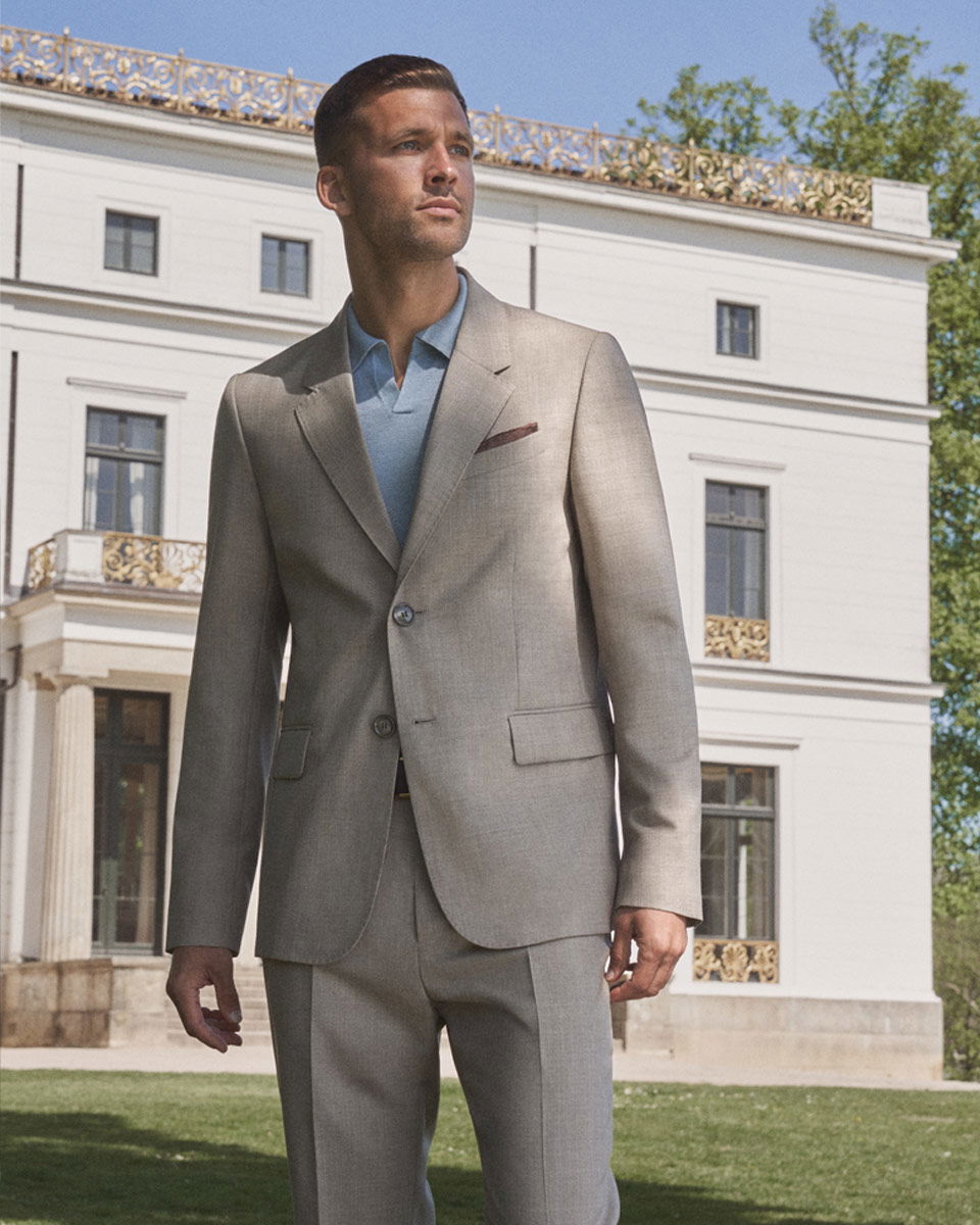 Mann im hellgrauen Anzug mit hellblauem Poloshirt vor weißem Herrenhaus bei Sonnenschein, Sommerlook.