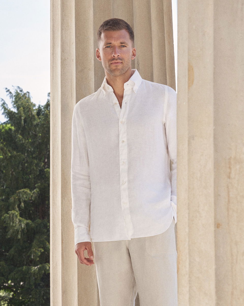 Mann in weißem Leinenhemd und cremefarbener Leinenhose steht vor hellen Säulen mit grünen Bäumen im Hintergrund.