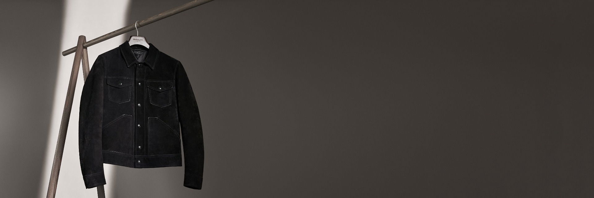 Schwarze Herren-Velourslederjacke mit Kragen und Taschen, hängend an einem Holz-Kleiderständer vor dunkelgrauem Hintergrund. Schwarze Herren-Velourslederjacke mit Kragen und Taschen, hängend an einem Holz-Kleiderständer vor dunkelgrauem Hintergrund.