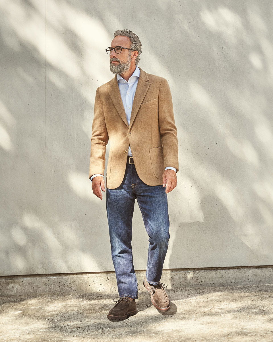 Mann mit grauem Haar und Bart trägt hellbraunes Sakko, hellblaues Hemd, dunkelblaue Jeans und braune Schnürschuhe vor heller Wand. Mann mit grauem Haar und Bart trägt hellbraunes Sakko, hellblaues Hemd, dunkelblaue Jeans und braune Schnürschuhe vor heller Wand.