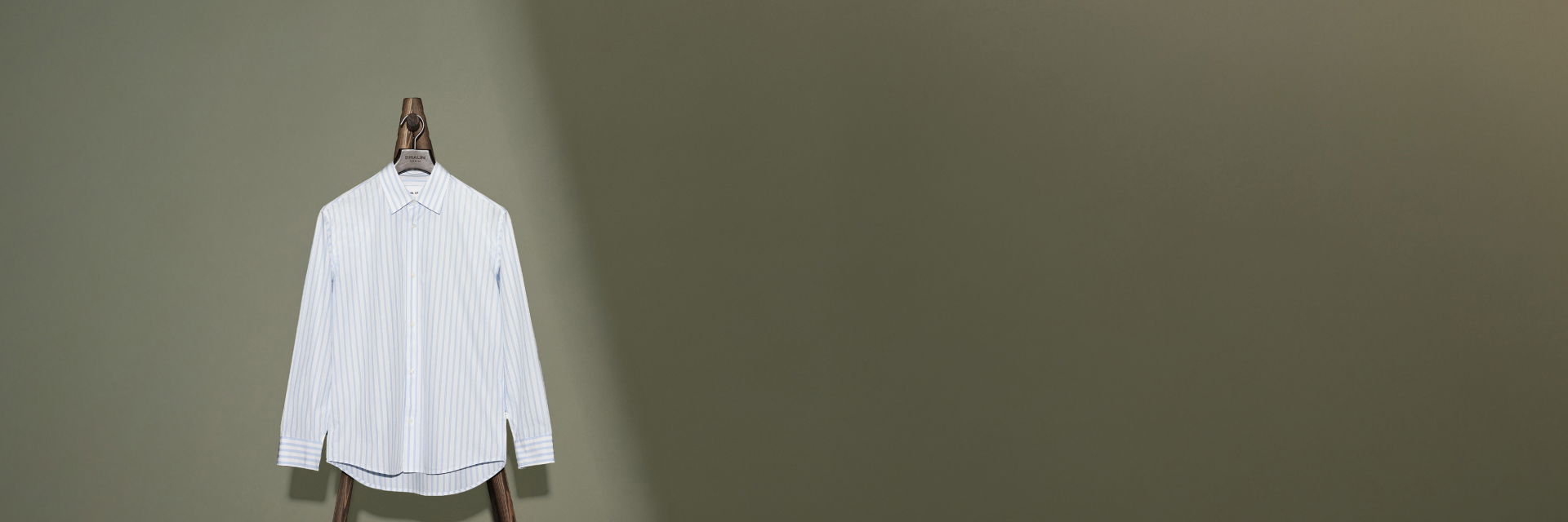Weiß-blau gestreiftes Herrenhemd auf Holzbügel vor olivgrünem Hintergrund hängend, Knopfleiste sichtbar. Weiß-blau gestreiftes Herrenhemd auf Holzbügel vor olivgrünem Hintergrund hängend, Knopfleiste sichtbar.