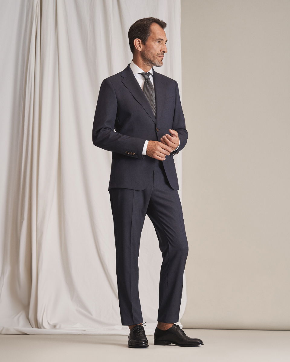 Mann im navy Anzug mit weißem Hemd, dunkelgrauer Krawatte und schwarzen Schnürschuhen vor hellem Hintergrund.
