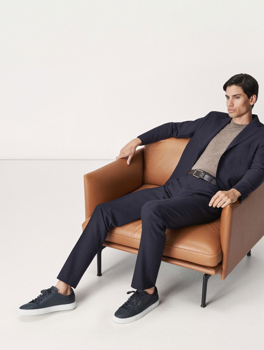 Mann sitzt entspannt in braunem Ledersessel, trägt navy Anzug, braunen Strickpullover und dunkelblaue Sneaker mit weißer Sohle. Mann sitzt entspannt in braunem Ledersessel, trägt navy Anzug, braunen Strickpullover und dunkelblaue Sneaker mit weißer Sohle.