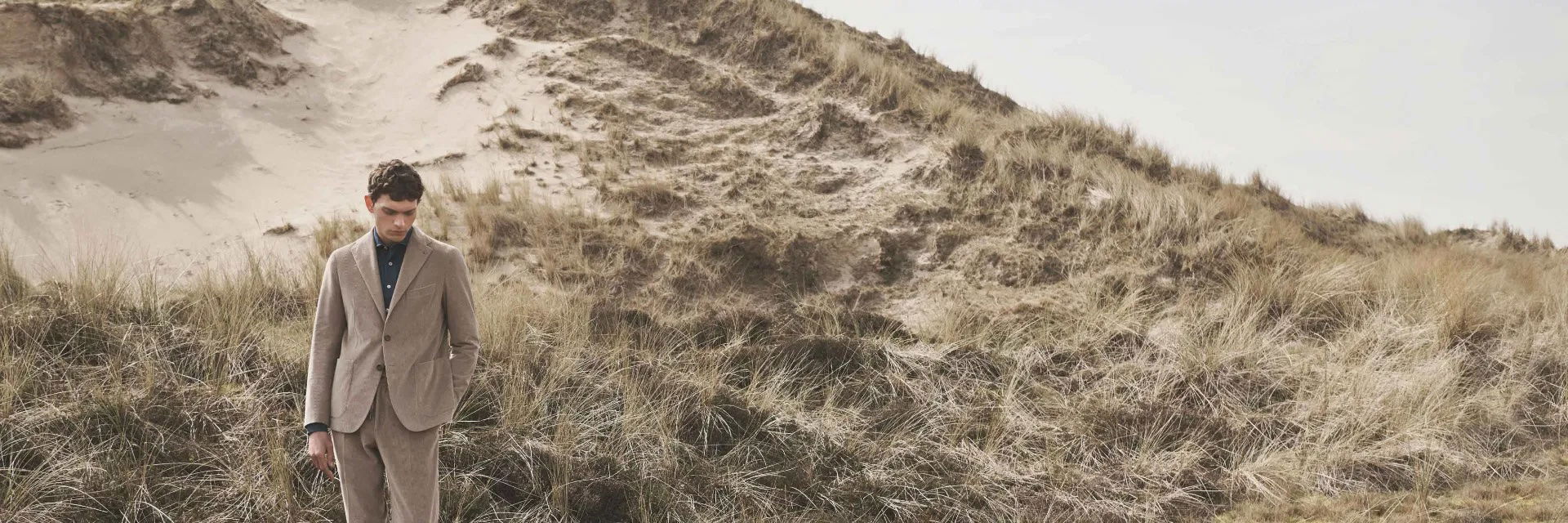 Mann in beigem Cordanzug steht nachdenklich in Dünenlandschaft mit trockenem Gras und sandigen Hügeln im Hintergrund. Mann in beigem Cordanzug steht nachdenklich in Dünenlandschaft mit trockenem Gras und sandigen Hügeln im Hintergrund.