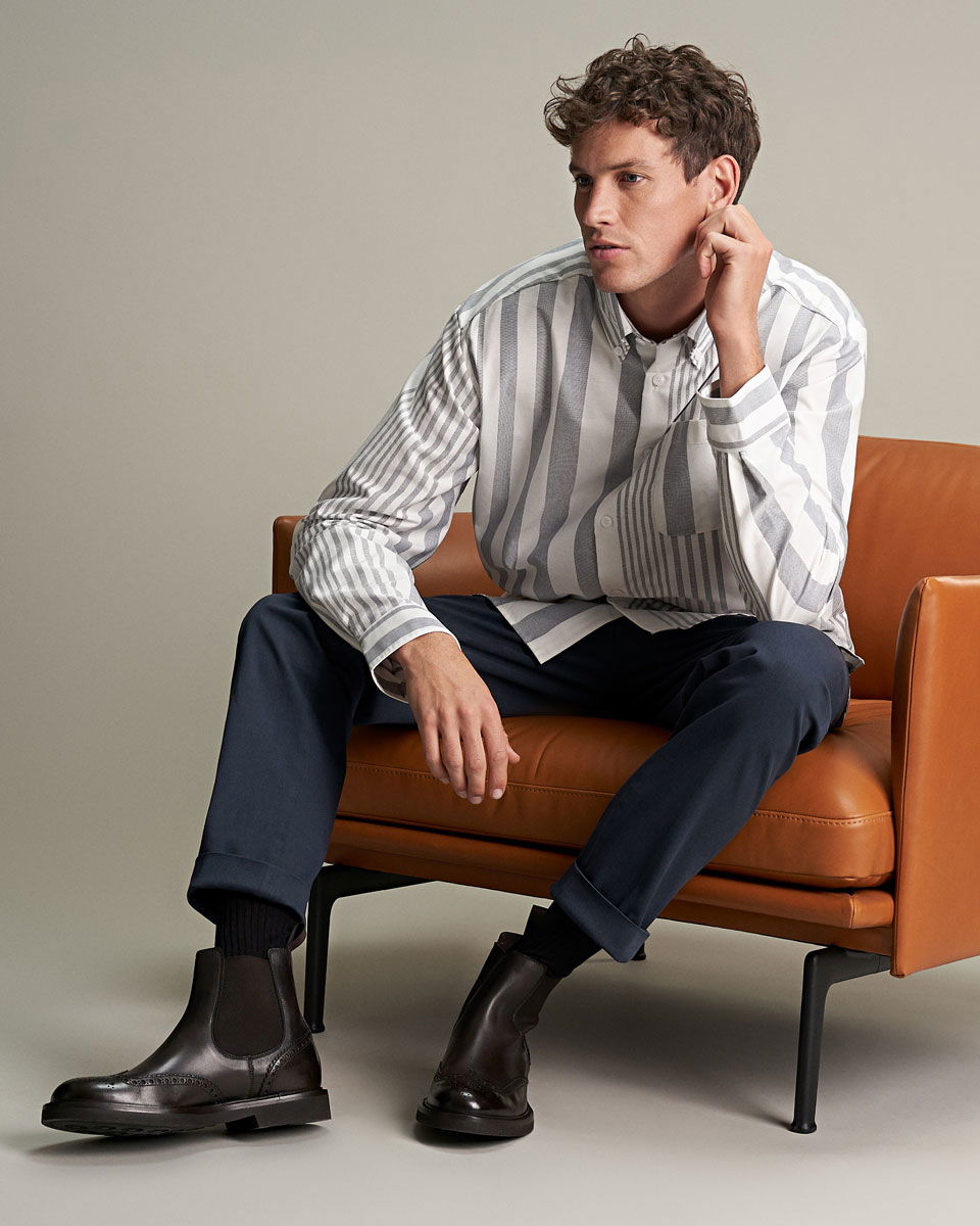 Mann sitzt auf braunem Ledersessel, trägt grau-weiß gestreiftes Hemd, dunkelblaue Hose und schwarze Chelsea Boots. Mann sitzt auf braunem Ledersessel, trägt grau-weiß gestreiftes Hemd, dunkelblaue Hose und schwarze Chelsea Boots.